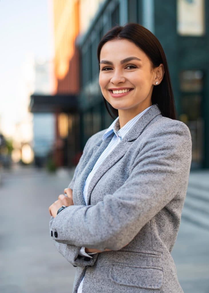 A smiling business woman in a suit standing in front of a building.