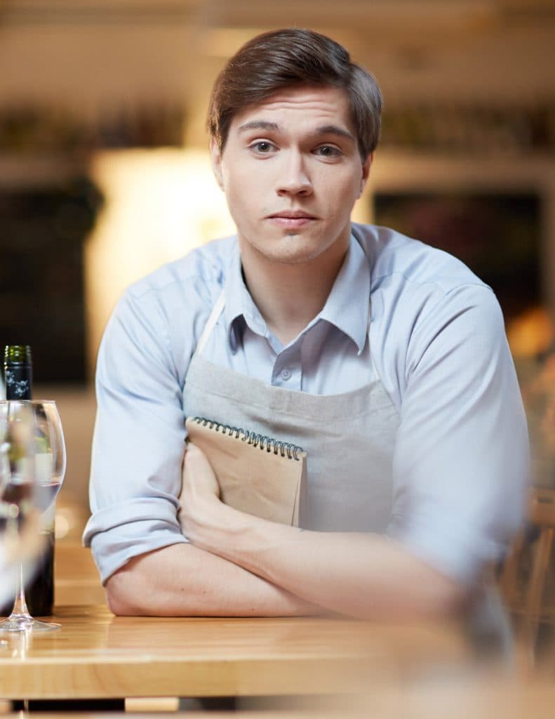 A man in a blue apron sitting at a table with a bottle of wine.