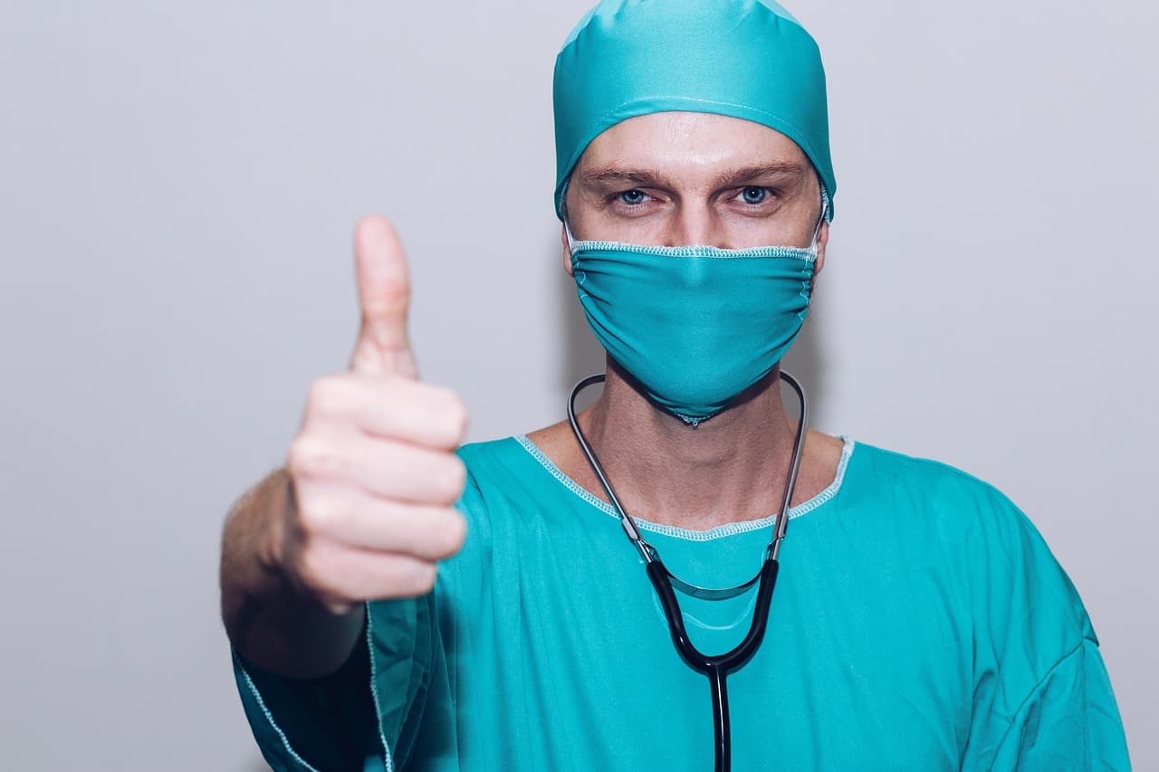 A healthcare professional in teal scrubs, cap, and mask gives a thumbs-up. A stethoscope hangs around their neck.