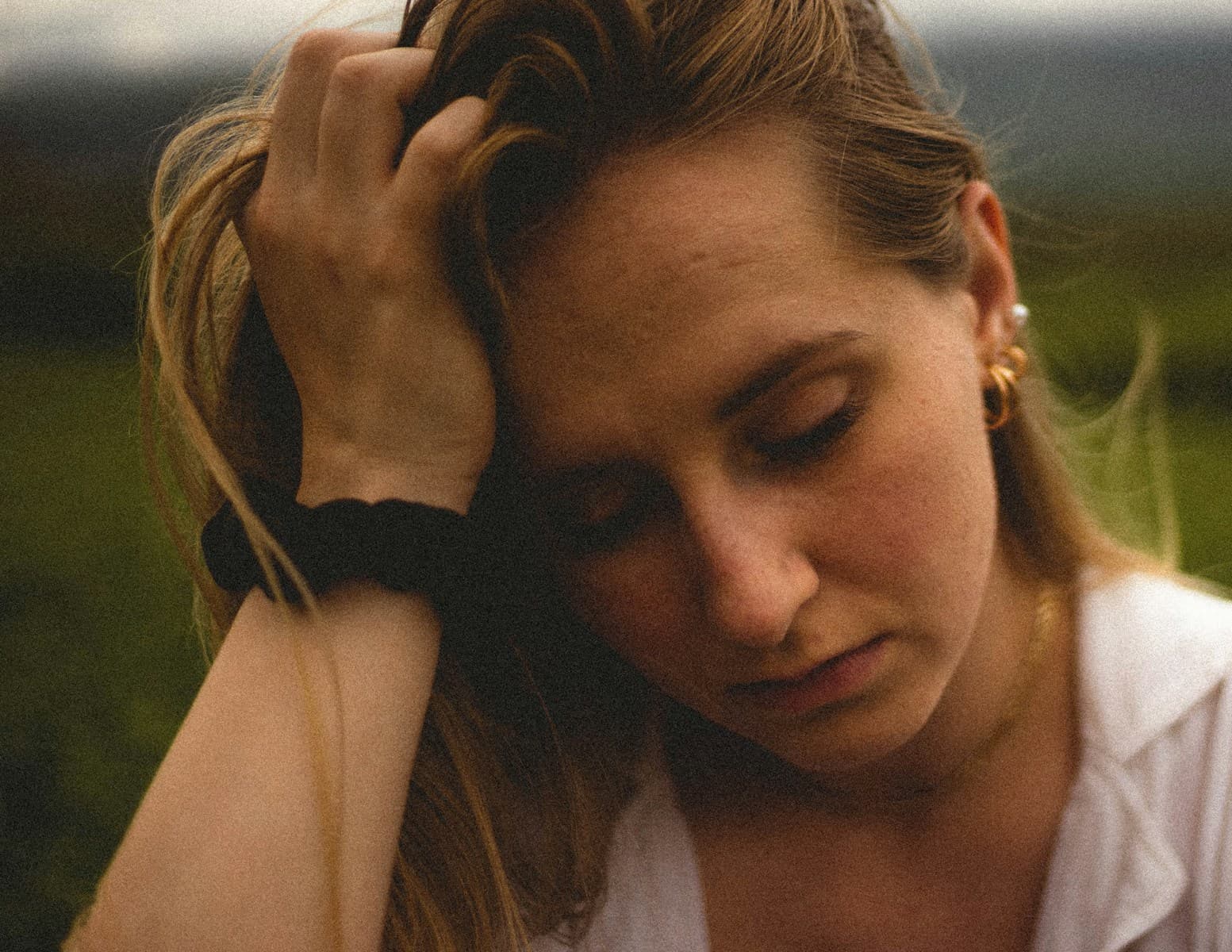 A person with closed eyes and a contemplative expression holds their head with one hand, wearing a white shirt and gold earrings, standing outdoors.