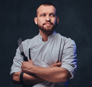 Um chef barbudo posando com os braços cruzados sobre um fundo escuro.