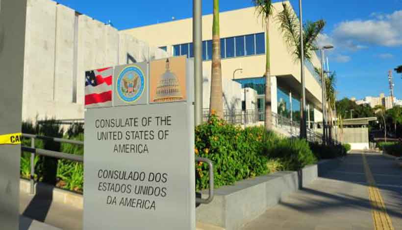 Placa dizendo "Consulado dos Estados Unidos da América" do lado de fora de um edifício moderno cercado por palmeiras. Bandeiras e um emblema são visíveis na placa.