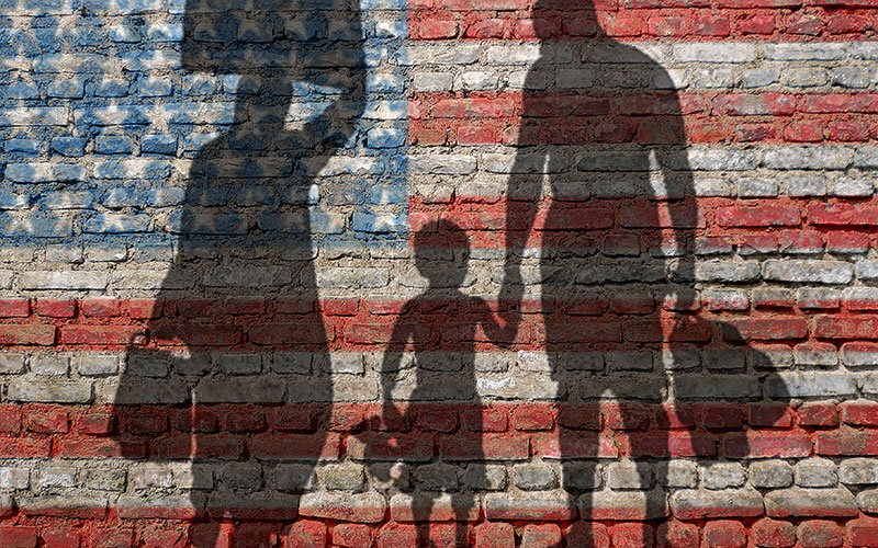 Sombras de três pessoas de mãos dadas carregando sacolas são projetadas em uma parede de tijolos pintada com a bandeira americana.