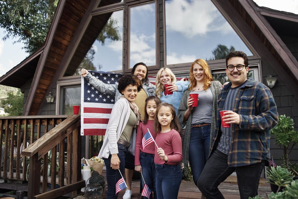 Um grupo de pessoas está em frente a uma casa com uma bandeira americana. Alguns estão segurando pequenas bandeiras e copos vermelhos. A casa tem um estilo de estrutura em A com janelas grandes.