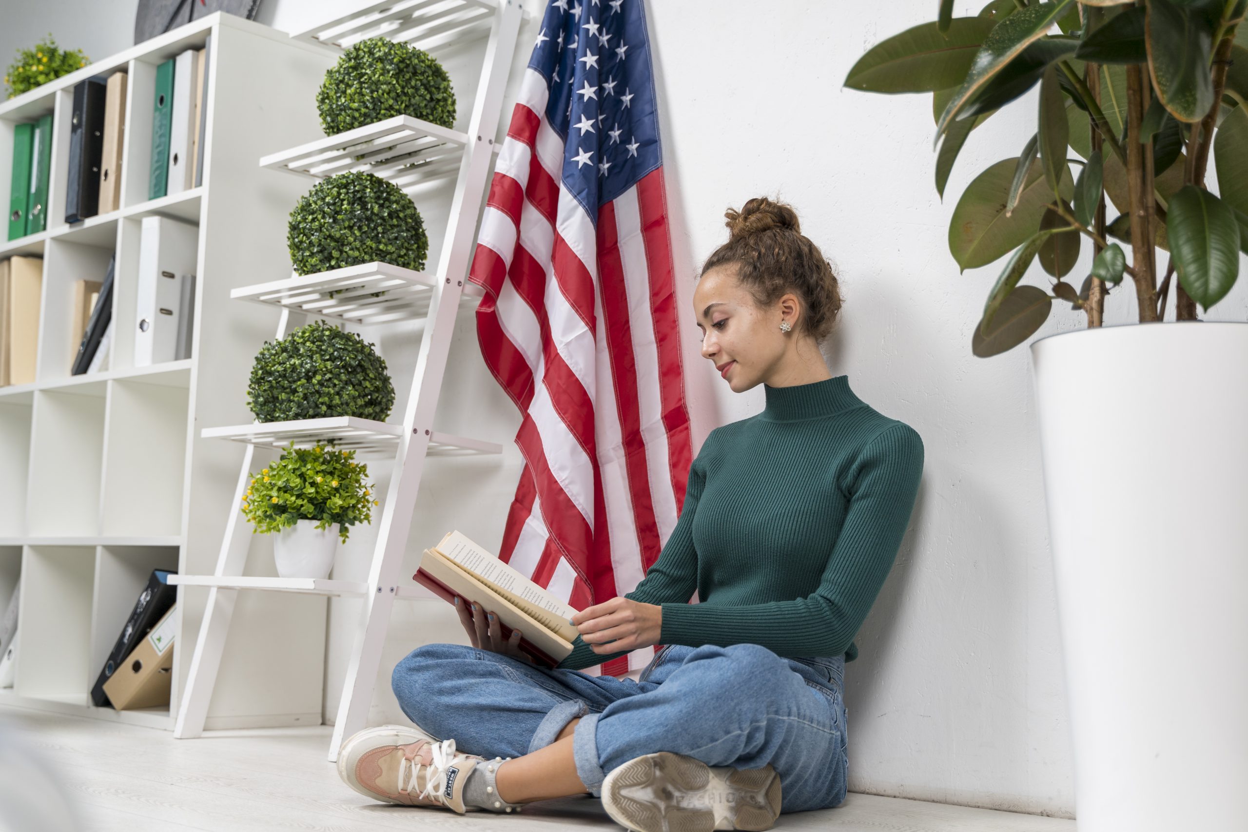 Mulher com uma gola alta verde sentada de pernas cruzadas no chão lendo um livro. Uma bandeira americana e plantas verdes em vasos em uma prateleira estilo escada estão ao fundo.