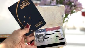 Uma pessoa segura um passaporte brasileiro e um passaporte aberto exibindo uma página de visto com uma foto de uma criança e detalhes do visto.