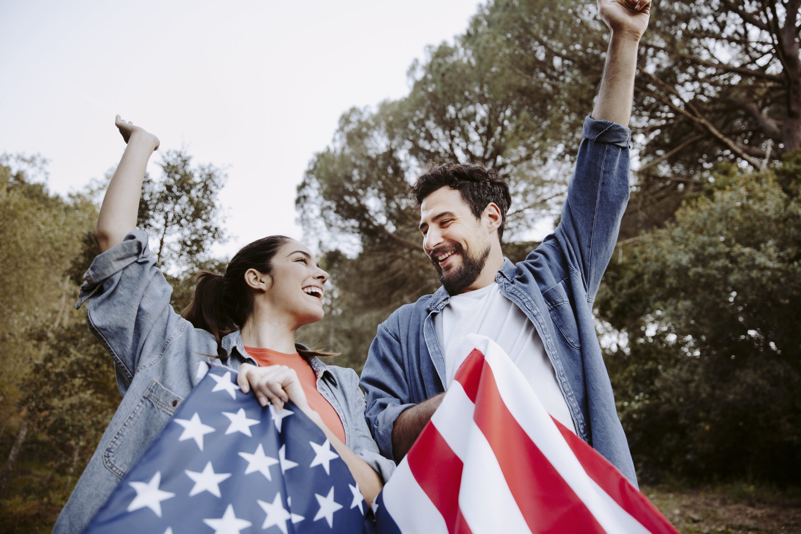 Um homem e uma mulher sorriem e seguram uma bandeira americana ao ar livre, ambos vestindo jaquetas jeans e levantando um braço no ar. Árvores são visíveis ao fundo.