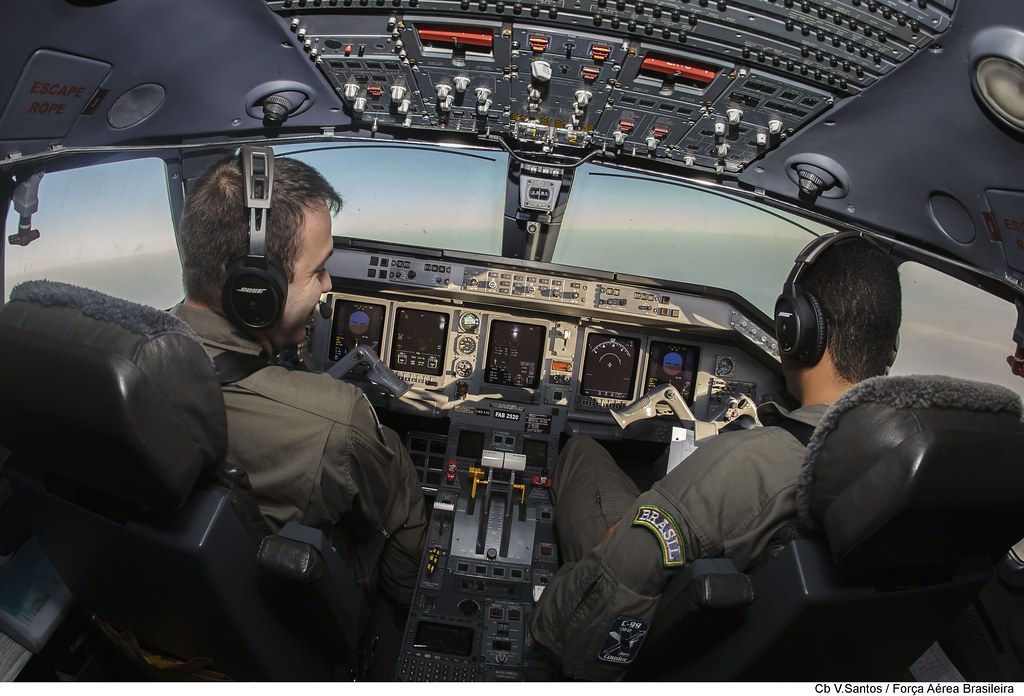 Dois pilotos em uniformes militares sentam-se na cabine de um avião. Eles estão usando fones de ouvido e olhando para o painel de controle e instrumentos do avião. A vista através das janelas da cabine mostra o céu e o horizonte.